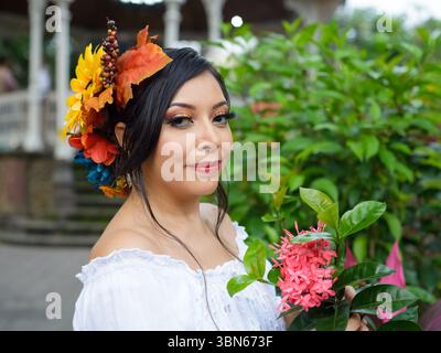Eine junge Frau mit dunklem Haar und herbstlichen Blumen hält eine leuchtende rosa Blüte. Sie trägt ein weißes schulterfreies Oberteil. Stockfoto