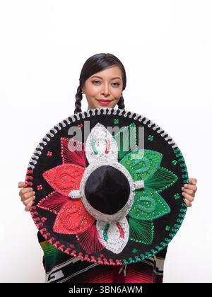 Eine junge Frau mit dunklen Haaren in Zöpfen lächelt sanft und hält einen großen, farbenfrohen mexikanischen Sombrero Stockfoto
