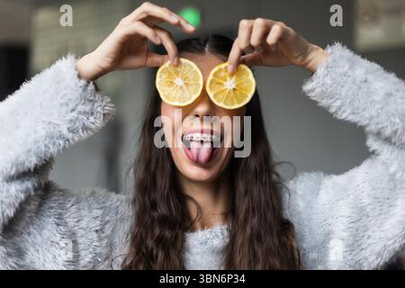 Die verspielte junge Frau lächelt mit einer Zahnspange und hält Zitronenscheiben an ihre Augen und drückt Freude und Kreativität in einer lässigen und unterhaltsamen Umgebung aus. Stockfoto