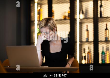 In einer eleganten Bar drückt eine junge Frau Müdigkeit oder Stress aus, während sie lange an ihrem Laptop arbeitet. Die Umwelt deutet auf eine Arbeitsszene außerhalb der Geschäftszeiten hin Stockfoto