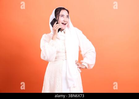 Eine attraktive muslimische Frau in weißem Kleid und Hijab, die glücklich auf ihrem Smartphone spricht und moderne Kommunikation und Lebensstil zeigt, isoliert auf einem Stockfoto