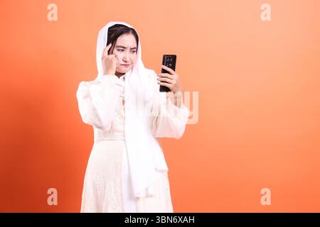 Junge muslimische Frau im Hijab, die verwirrt aussah, während sie ihr Smartphone benutzte und Unsicherheit und Kontemplation in moderner Kommunikation zeigte, isoliert auf Stockfoto
