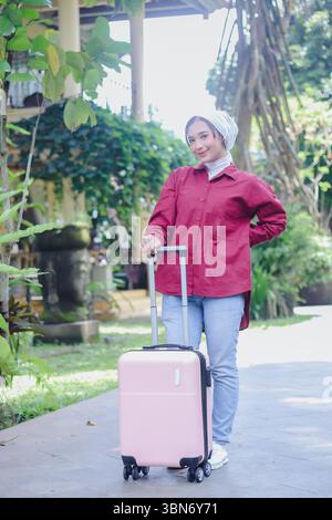 Eine hübsche muslimische Frau, die einen stilvollen Hijab trägt, zieht fröhlich einen rosafarbenen Koffer und ist bereit für eine aufregende Reise in einer reizvollen Umgebung Stockfoto
