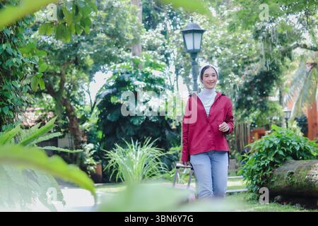 Eine strahlende junge Frau, die einen Hijab trägt und sich freudig auf ihr Reiseabenteuer einlässt. Sie zieht einen Koffer in der ruhigen Schönheit eines üppigen Gartens Stockfoto