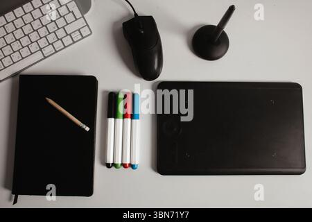 Moderner Arbeitsbereich mit Tastatur, schwarzem Notebook, Bleistift, Grafiktablett und Stiften auf weißem Schreibtisch. Draufsicht für kreatives Projekt. Stockfoto