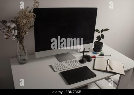 Minimalistischer Home Office-Arbeitsplatz mit Desktop-Computer, Tastatur, Notebook, Stifttablett, getrockneten Blumen und einer Pflanze auf einem sauberen weißen Schreibtisch. Arbeiten Sie zu Hause Stockfoto