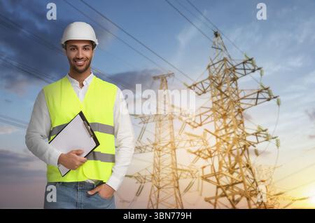 Der Techniker im Schutzhelm hält das Klemmbrett gegen Hochspannungsmasten Stockfoto