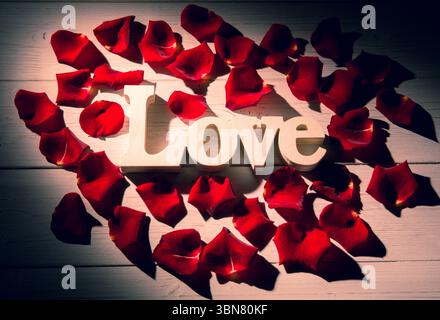 Der Love-Ausschnitt aus Holz liegt auf einer flachen Oberfläche aus Illustrationsholz, mit roten Rosenblättern, die das Herz bilden Stockfoto