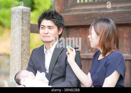 Im Juni 2025 genießt ein junges japanisches Paar Anfang 20 die omiyamairi ihres Babys, indem es auf einer Steinmauer an einem Schrein in Aichi sitzt, friedlich Stockfoto