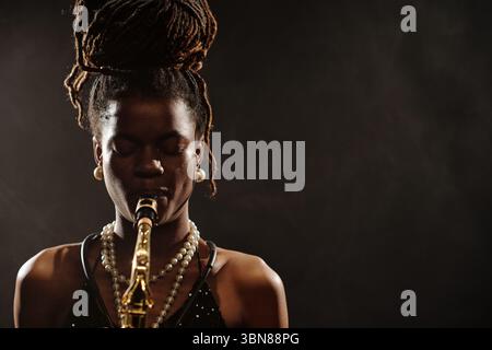 Schwarze junge Frau spielt Saxophon mit geschlossenen Augen, trägt Perlenkette und Ohrringe, steht vor dunklem Hintergrund und verkörpert die Aufführung eines Jazzmusikers Stockfoto