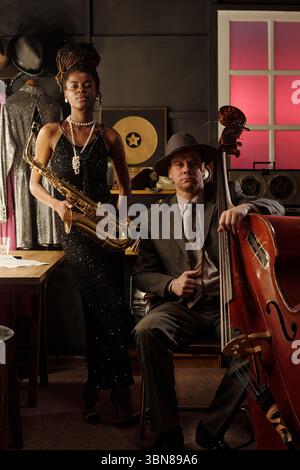 Potrait einer schwarzen jungen erwachsenen Frau mit Saxophon und sitzendem kaukasischen Mann mittleren Alters mit Kontrabass in Vintage-Musik-Studio-Setting, mit Jazz-Erinnerungsstücken sichtbar Stockfoto