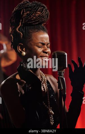 Schwarze junge Erwachsene Frau, die leidenschaftlich in ein Vintage-Mikrofon auf der Bühne singt, die Augen geschlossen, elegante Accessoires trägt und Jazzmusik mit aufrechten Bässen im Hintergrund vorführt Stockfoto