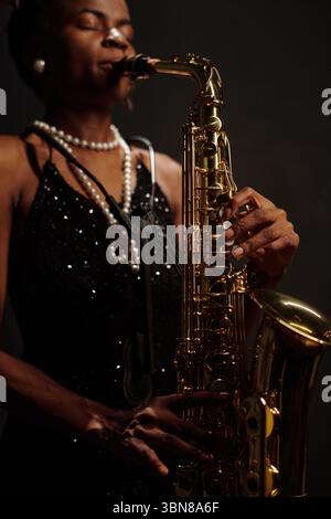 Afroamerikanische Musikerin, die Saxophon auf der Bühne spielt, Schmuck trägt und Jazzmusik aufführt, Hände packend, Augen geschlossen, Schwerpunkt auf musikalischem Ausdruck Stockfoto