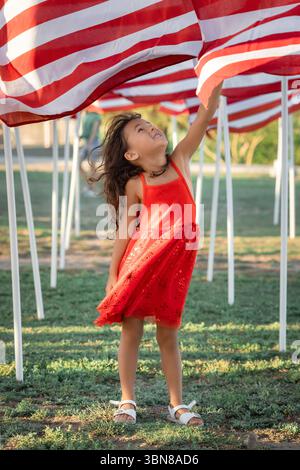 Lächelndes Mädchen in rotem Kleid, das am Memorial Day auf die amerikanische Flagge blickt, patriotische Kinder und Nationalfeierlichkeiten – Bild Stockfoto