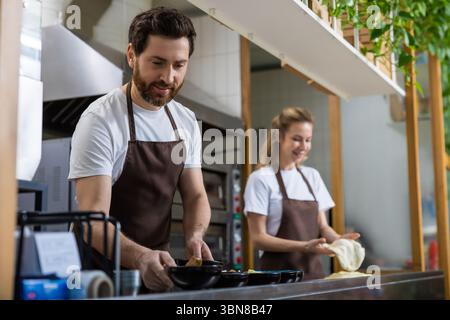 Mann und Frau mit Schürzen arbeiten nebeneinander in einer gut beleuchteten Bäckereiküche Stockfoto