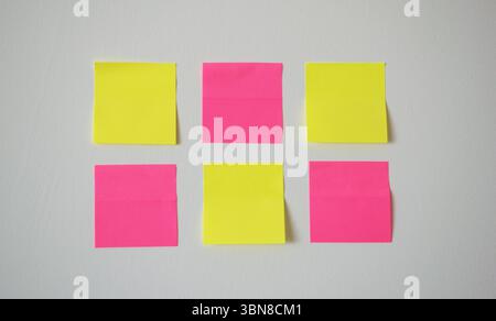 Minimales und kreatives Arrangement von sechs leeren Haftnotizen in Nahaufnahme, drei gelben und drei rosa, ordentlich auf einer hellen weißen Wand platziert. Stockfoto