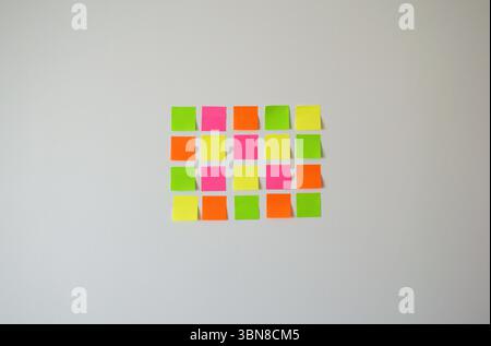 Organisiertes Raster aus zwanzig bunten Haftnotizen aus nächster Nähe, Rosa, Gelb, Orange und Grün, ordentlich auf einer hellweißen Wand angeordnet. Minimal und sauber. Stockfoto