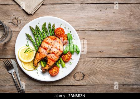 Gegrilltes Lachssteak mit grünem Spargel, Tomaten und Zitronenscheiben auf hölzernem Hintergrund, Blick von oben, Kopierraum. Ausgewogene gesunde Mahlzeit, Keto-Mittagessen. Stockfoto