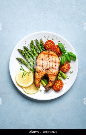 Gegrilltes Lachssteak mit grünem Spargel, Tomaten und Zitronenscheiben auf weißem Teller und blauem Hintergrund, Draufsicht. Ausgewogene gesunde Mahlzeit, Keto-Mittagessen. Stockfoto