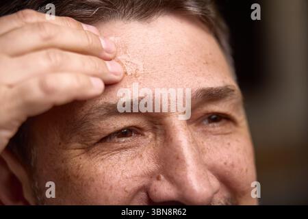 Schönheit und Alterung. Reifer Mann, der Hautpflegemittel auf die Stirn aufträgt, Nahaufnahme Stockfoto