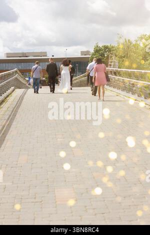 An einem teilweise bewölkten Tag läuft eine Gruppe von Menschen über eine sonnendurchflutete Fußgängerbrücke. Die Hochzeitszeremonie geht in die Ferne entlang der Straße, eine Ehe Stockfoto