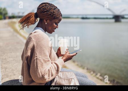Die junge schwarze Frau benutzt Smartphone und trinkt Kaffee, um draußen am Fluss zu gehen. Stockfoto
