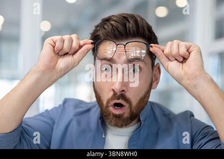 Ein Mann mit Brille ist überrascht, hält seine Brille mit einem Blick von Erstaunen und Unglauben. Stockfoto