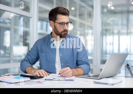 Ein Mann in einem blauen Hemd sitzt an einem Schreibtisch und schreibt auf Papier. Ein Laptop, ein Taschenrechner und andere Büromaterialien befinden sich auf dem Schreibtisch. Stockfoto