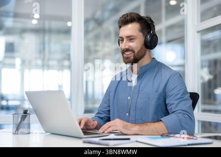 Ein lächelnder Mann mit Kopfhörern arbeitet in einem modernen Büro an einem Laptop und sieht glücklich aus. Stockfoto