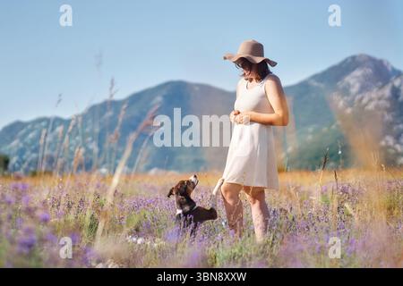 Ein kleiner Hund steht auf einer Lavendelwiese neben einem Mädchen, das ein weißes Kleid und einen breitkrempigen Hut trägt. Das sanfte Licht hebt die leuchtenden Blumen und den hervor Stockfoto