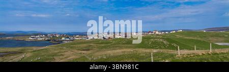 Ein Panoramablick auf die Küstengemeinde Hamnavoe in Shetland, Großbritannien, eingebettet zwischen sanften grünen Hügeln und einer ruhigen Bucht unter einem klaren blauen Himmel. Stockfoto