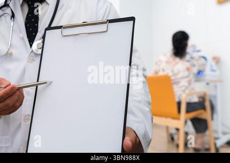 Nahaufnahme eines Arztes, der ein leeres Klemmbrett mit Stift präsentiert, bereit für Notizen oder Diagnose in einer medizinischen Klinik. Eine Patientin wird im Hintergrund gesehen Stockfoto