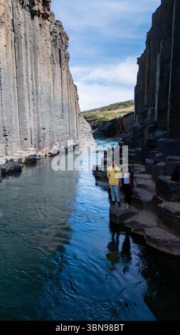 Besucher bestaunen die atemberaubende Schönheit des Studlagil Canyon, wo sich majestätische Basaltsäulen dramatisch über kristallklarem Wasser erheben. Dieses geologische Wunder zeigt die einzigartige Landschaft Islands. Stockfoto