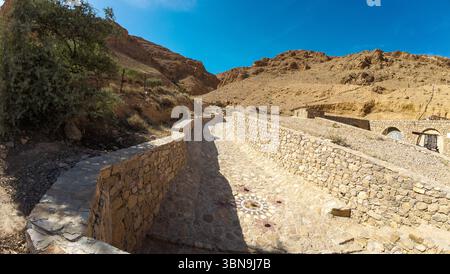Ein Steinweg mit Mosaikmotiven schlängelt sich durch eine felsige Wüstenlandschaft unter einem klaren blauen Himmel. Stockfoto