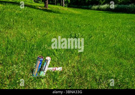Drei gebrauchte Feuerwerkskannister im Gras eines Parks mit Bäumen im Hintergrund an einem sonnigen Tag Stockfoto