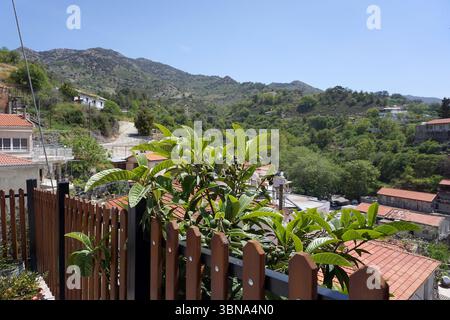 Eine ruhige Berglandschaft mit einem klaren blauen Himmel. Die Berge sind von üppiger grüner Vegetation bedeckt und das Gelände ist hauptsächlich felsig und hügelig. Im Vordergrund steht ein hölzerner Zaun mit einem schwarzen Metallgeländer, auf dem ein kleiner Baum oder Büsche wächst. Hinter dem Zaun befindet sich eine kleine Siedlung im Tal mit Gebäuden mit roten Ziegeldächern. Die Siedlung ist von einer Mischung aus Bäumen und Vegetation umgeben, und in der Mitte ist eine asphaltierte Straße oder ein gepflasterter Pfad zu sehen. Das Bild wird von einem hohen Aussichtspunkt aufgenommen und bietet einen Panoramablick auf die Siedlung und den umliegenden Moun Stockfoto