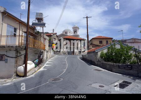 Zypern Dorf, Eine enge, gewundene Straße. Die Straße ist von alten Steinhäusern gesäumt, von denen einige rote Ziegeldächer haben und einige Bäume und Büsche aufweisen. Im Hintergrund ist eine Kirche mit einer weißen Kuppel und einem Glockenturm zu sehen. An der Straße befinden sich Versorgungsmasten und Stromleitungen, und auf der linken Seite ist ein Wassertank zu sehen. Die Straße scheint eine Einbahnstraße zu sein, und auf der rechten Seite ist ein Schild mit der Aufschrift „Taverna“ zu sehen. Der Himmel ist ein klares Blau mit ein paar Stockfoto