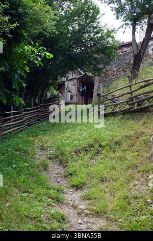 Bulgarien, Gela, Eine rustikale Szene in ländlicher Umgebung. Ein unebener Pfad schlängelt sich durch einen grasbewachsenen Bereich und führt zu einer Holzstruktur, die einer Scheune oder einem Schuppen ähnelt. Die Struktur wird teilweise durch eine Steinmauer und einen Holzzaun aus Holzpfosten und -Schienen verdeckt. Die Gegend ist von üppigem Grün umgeben, mit Bäumen und Büschen, die die Landschaft prägen. Der Himmel ist hellblau und der Gesamteindruck ist von Ruhe und natürlicher Schönheit. Das Auge und die Phantasie eines Künstlers Stockfoto