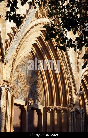 Zypern, Famagusta, Kathedrale des Heiligen Nikolaus, Lala Mustafa Pascha Moschee, mittelalterliche Kirche, die 1571 von den Türken in eine Moschee umgewandelt wurde, mit einem Minarett mit Blick auf das Meer., Eine gotische Kathedrale mit einem großen, reich verzierten Bogengang, der die Szene dominiert. Der Bogengang ist aus hellem Stein gefertigt und mit komplizierten Schnitzereien und Mustern versehen, darunter ein Rosenfenster mit einem detaillierten Blumendesign. Die Fassade der Kathedrale besteht hauptsächlich aus hellem Stein mit einigen dunkleren Steinelementen, und der Bogengang wird von zwei dunkelbraunen Holztüren flankiert. Die Perspektive des Bildes ist von unten. Stockfoto