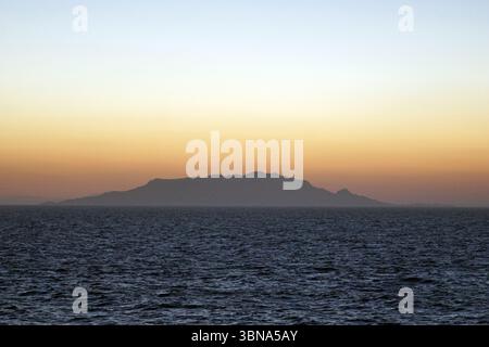 Griechische Inseln zwischen Zypern und Athen. Ein ruhiger Sonnenuntergang über einer riesigen Weite des dunkelblauen Ozeans. Der Himmel ist in Orange- und Gelbtönen gemalt, während die Sonne hinter einer Bergkette in der Ferne untergeht. Die Bergkette, die sich vor dem pulsierenden Himmel bildet, scheint eine große, möglicherweise vulkanische Landmasse zu sein. Die Oberfläche des Ozeans ist relativ ruhig und weist kleine Wellen auf. Das Bild wird aus der Ferne aufgenommen und bietet eine weite Sicht auf die Landschaft und den Horizont. Das Auge und die Phantasie eines Künstlers Stockfoto