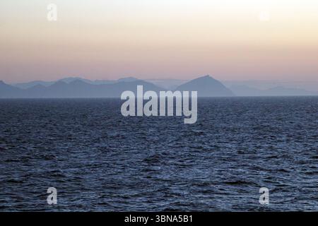 Griechische Inseln zwischen Zypern und Athen. Eine ruhige Küstenlandschaft bei Sonnenaufgang oder Sonnenuntergang. Der Himmel ist in Rosa- und Orange-Tönen gemalt und strahlt ein warmes Leuchten über die Szene. Der ruhige, dunkelblaue Ozean erstreckt sich bis zum Horizont, wo sich in der Ferne eine Reihe von Bergen, hauptsächlich dunkelgrau oder bläulich-grau, erheben. Die Berge wirken trüb oder nebelig und verleihen der Szene ein Gefühl von Tiefe und Geheimnis. Das Bild wurde aus der Ferne aufgenommen und bietet einen Panoramablick auf die Landschaft. Das Auge und die Phantasie eines Künstlers Stockfoto