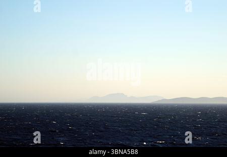 Griechische Inseln zwischen Zypern und Athen. Ein riesiger, dunkelblauer Ozean, der sich bis zum Horizont erstreckt. Die Horizontlinie ist deutlich sichtbar und trennt den Ozean vom Himmel. Der Himmel hat eine hellblaue Farbe mit einem trüben oder nebeligen Aussehen in der Entfernung. Die Horizontlinie ist nicht perfekt gerade, mit einer leichten Kurve. In der Ferne kann man eine Reihe von trüben, hellen Bergen sehen. Die Berge sind nicht scharf definiert und erscheinen als trübe Silhouette am Himmel. Das Auge und die Phantasie eines Künstlers Stockfoto