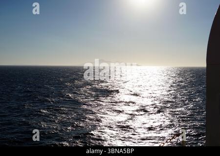 Griechische Inseln zwischen Zypern und Athen. Ein ruhiger Blick auf das Meer. Das tiefblaue Wasser ist mit kleinen Wellen gespickt, die das Sonnenlicht reflektieren und einen schimmernden Effekt auf der Oberfläche erzeugen. In der Ferne ist eine schwache Silhouette einer Landmasse, möglicherweise eines Gebirges, vor dem klaren, hellblauen Himmel zu sehen. Die Sonne befindet sich in der oberen rechten Ecke und strahlt ein helles Licht aus, das die Szene beleuchtet. Ein Teil des Geländes oder Zauns eines Schiffes ist in der unteren rechten Ecke sichtbar, was darauf hindeutet, dass die Perspektive vom Deck aus ist. Das Auge und die Phantasie eines Künstlers Stockfoto