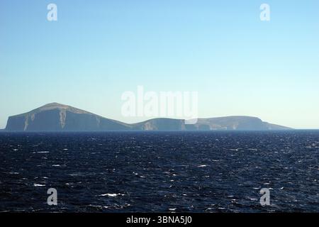 Griechische Inseln zwischen Zypern und Athen. Eine ruhige Szene eines riesigen, dunkelblauen Ozeans, das auf eine hellbraune, felsige Küste trifft. Der Himmel ist klar, hellblau und bietet einen schönen Kontrast zum tiefen Blau des Ozeans. Die Küste besteht aus einer Reihe von felsigen, dunkelbraunen Klippen, die sich aus dem Wasser erheben und in der Ferne einige kleinere, weniger definierte Inseln zu sehen sind. Das Bild wurde aus der Ferne aufgenommen und bietet einen Panoramablick auf das Meer und die Küste, ohne sichtbare Boote oder Schiffe. Das Auge und die Phantasie eines Künstlers Stockfoto