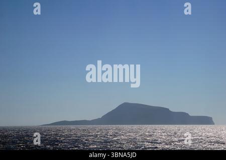 Griechische Inseln zwischen Zypern und Athen. Eine ruhige Szene einer dunklen, möglicherweise vulkanischen Insel inmitten eines riesigen, dunkelblauen Ozeans. Die Insel hat einen kleinen, abgerundeten Gipfel und ist vom Meer umgeben, das eine leicht abgehackte Oberfläche hat. Der Himmel darüber ist klar, hellblau, ohne Wolken. Das Bild wurde aus der Ferne aufgenommen und bietet einen umfassenden Blick auf die Insel und das Meer. Das Auge und die Phantasie eines Künstlers Stockfoto