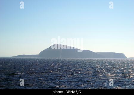 Griechische Inseln zwischen Zypern und Athen. Eine ruhige Küstenlandschaft mit einer großen, dunkel gefärbten, felsigen Insel im Zentrum. Die Insel hat einen markanten, runden Gipfel und scheint von Vegetation bedeckt zu sein. Der tiefblaue Ozean erstreckt sich bis zum Horizont, mit kleinen Wellen, die sanft gegen das Ufer plätschern. Der Himmel ist klar, hellblau, und die Horizontlinie ist in der Entfernung sichtbar. Das Bild wurde aus der Ferne aufgenommen und bietet einen weiten Blick auf die Insel und das Meer. Das Auge und die Phantasie eines Künstlers Stockfoto