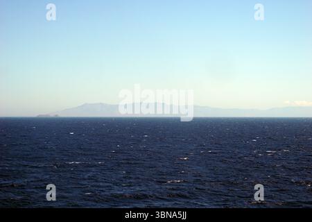 Griechische Inseln zwischen Zypern und Athen. Ein riesiger, tiefblauer Ozean, der sich bis zum Horizont erstreckt. Der Himmel ist klar, hellblau, mit ein paar Wolken am Horizont. In der Ferne erhebt sich eine Reihe von Bergen, vor allem braun und braun, am Horizont. Die Berge scheinen eine Mischung aus Felsen und möglicherweise etwas Vegetation zu sein. Das Bild wird von einem hohen Aussichtspunkt aufgenommen und bietet einen Panoramablick auf das Meer und die fernen Berge. Das Auge und die Phantasie eines Künstlers Stockfoto