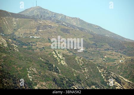 Griechische Inseln zwischen Zypern und Athen. Eine bergige Landschaft mit einem markanten, felsigen Gipfel im Hintergrund. Der Berg ist von einer Mischung aus grüner Vegetation und braunen oder braunen Flecken bedeckt, wobei einige Bereiche Anzeichen von Erosion oder Witterung zeigen. Die Perspektive ist von einem unteren Aussichtspunkt aus, der auf den Berg blickt und ein Gefühl von Größe und Pracht vermittelt. Der Himmel ist klar, hellblau, und einige kleine Strukturen, möglicherweise Häuser oder Gebäude, sind über die Landschaft verstreut. Auf dem Berggipfel ist auch ein Funkturm zu sehen. Das Auge und die Phantasie eines Künstlers Stockfoto