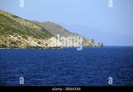 Griechische Inseln zwischen Zypern und Athen. Eine felsige Küste mit einem tiefblauen Meer im Vordergrund. Die Küste ist durch eine Mischung aus hellbraunen und braunen Felsen mit einigen Grünflächen gekennzeichnet. Das Meer wirkt relativ ruhig mit kleinen Wellen. In der Ferne erstreckt sich eine Reihe von Bergen über den Horizont, deren Gipfel von einem trüben blauen Himmel verdeckt werden. Die Berge sind eine Mischung aus braunen und braunen Farben. Das Auge und die Phantasie eines Künstlers Stockfoto