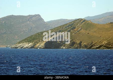 Griechische Inseln zwischen Zypern und Athen. Eine felsige Küste mit einem tiefblauen Meer im Vordergrund. Die Küste ist geprägt von steilen, felsigen Klippen und Hügeln mit einer Mischung aus grüner Vegetation und braunen oder braunen Flecken. Das Meer ist relativ ruhig mit kleinen Wellen. In der Ferne ist eine Windturbine auf einem Hügel sichtbar. Der Himmel ist klar, hellblau. Das Auge und die Phantasie eines Künstlers Stockfoto